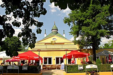 glebes Gebäude mit großen Laubbäumen und Gastgarten mit Sonnenschirmen im Vordergrund