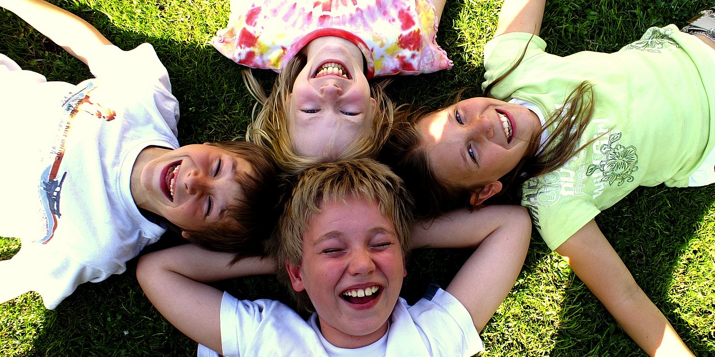 Vier Kinder, die mit ihren Köpfen an- und nebeneinander einen Kreis im Gras bilden 