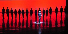 Tanzcompany, auf in rotes Licht getauchter Bühne, mit Choreograf im Vordergrund. 