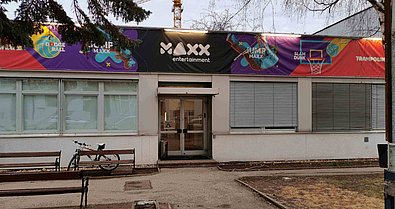 Die bunte Vorderseite eines Gebäude ist zu sehen; auf schwarz steht in weißer Schrift "MAXX". Neben dem Eingang steht ein Fahhrad hinter einer Sitzbank.