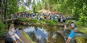 Grünes Waldstück mit einem kleinen See, bei dem einige Menschen sitzen.