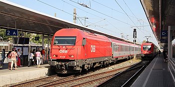 Bild von zwei Zügen, die am Bahnhof Wien Meidling stehen. Auf den Bahnsteigen sind Menschen, die einsteigen wollen oder auf einen nächsten Zug warten.