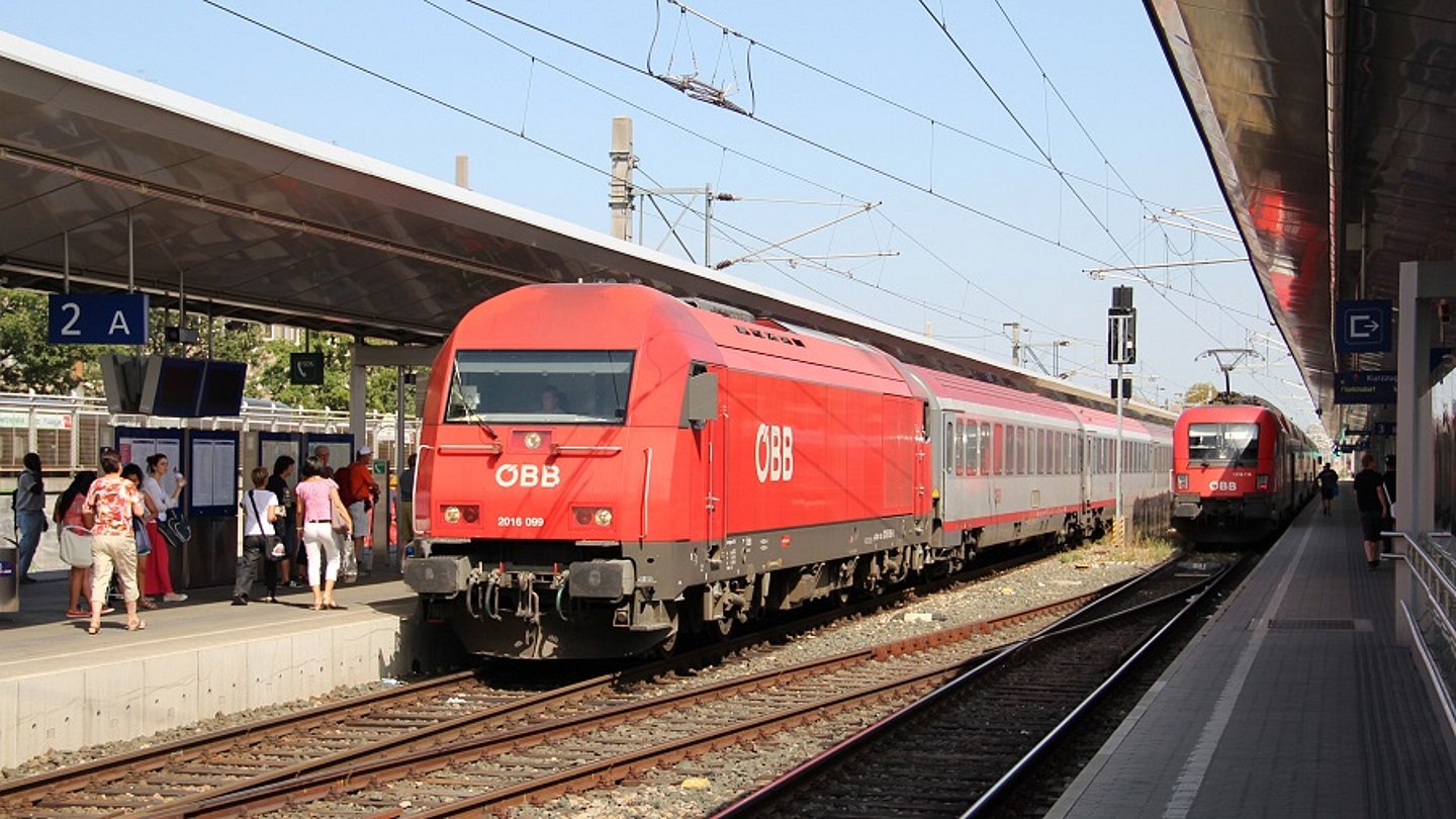 Bild von zwei Zügen, die am Bahnhof Wien Meidling stehen. Auf den Bahnsteigen sind Menschen, die einsteigen wollen oder auf einen nächsten Zug warten.