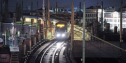 Bild der U-Bahn-Linie U6 bei Nachtbetrieb. Der Zug hat die Scheinwerfer an und ist in der Nähe der Station Längenfeldgasse unterwegs. Im Hintergrund befinden sich beleuchtete Häuser und die Gürtelbögen, sowie ein Basketballkorb und Graffitis.