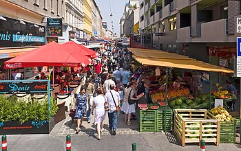 Bild vom Eingang des Brunnenmarktes: Lokale und Verkaufsstände auf beiden Seiten, letztere bieten frisches Obst und Gemüse an.