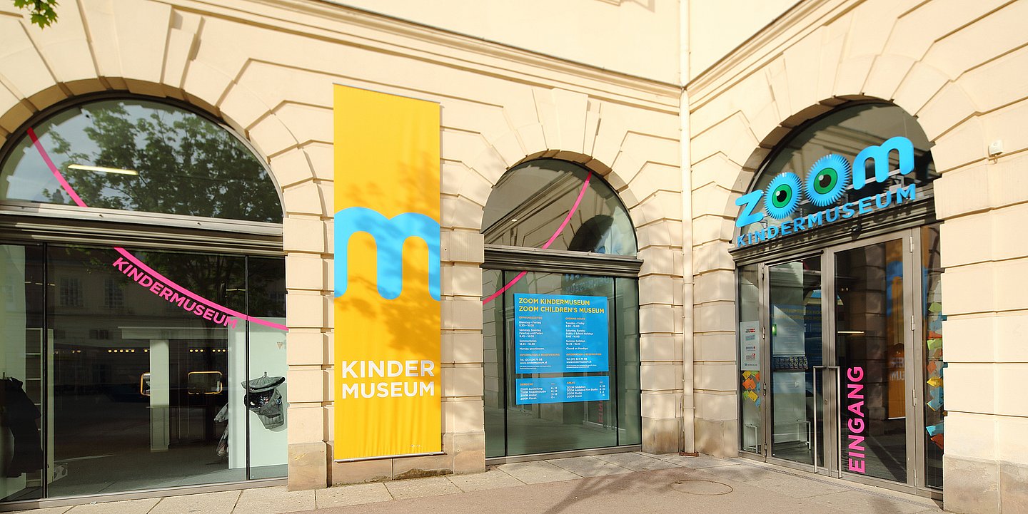 Außenansicht eines Gebäudes. Im Zentrum sind zwei Glastüren mit der Schrift ZOOM Kindermuseum.