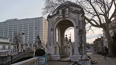 Ein großer Pavilion aus Stein ist zu sehen, während rechts von ihm ein blattloser Baum und links von ihm ein mehrstöckiges Gebäude zu sehen sind.