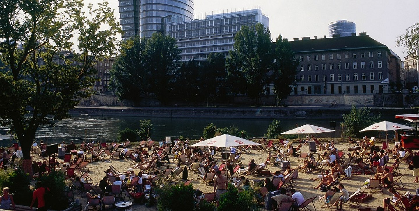 Viele Menschen sitzen auf Liegestühlen unter >Sonnenschirmen am Strand vorm Donaukanal