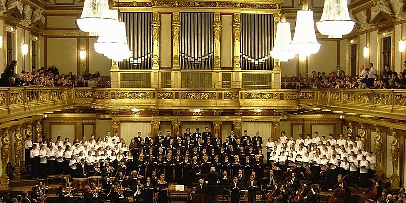 Orchesterkonzert mit Chor im Goldenen Saal im Wiener Musikverein. Publikum auf den Galerien, hell erleuchtete große Luster hängen von der Decke, im Hintergrund die goldene Orgel. 