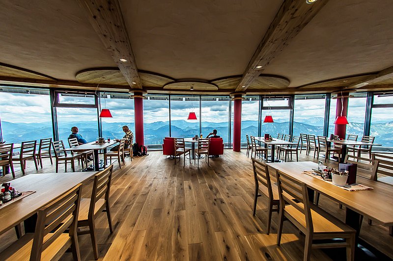 Tische, Sessel im Restaurant mit Fenster rundherum die eine breite Gebirgslandschaft zeigen